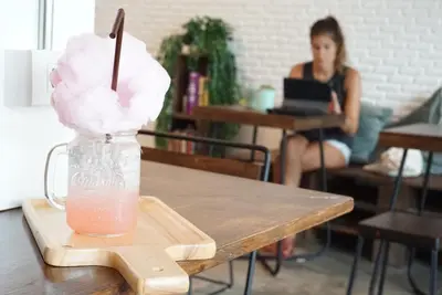 a drink on a wooden table with a woman working on a laptop at Krit Hostel in Bangkok