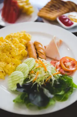 a white plate of food with eggs sausage and vegetables at Issara by D Hostel in Bangkok