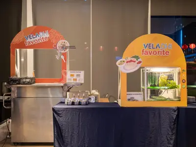 a table with a yellow and orange food stand next to a refrigerator at VELA Dhi GLOW Pratunam in Bangkok
