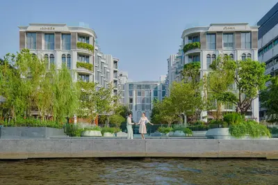two people standing on a sidewalk near the water with buildings at The Salil Hotel Riverside Bangkok in Bangkok